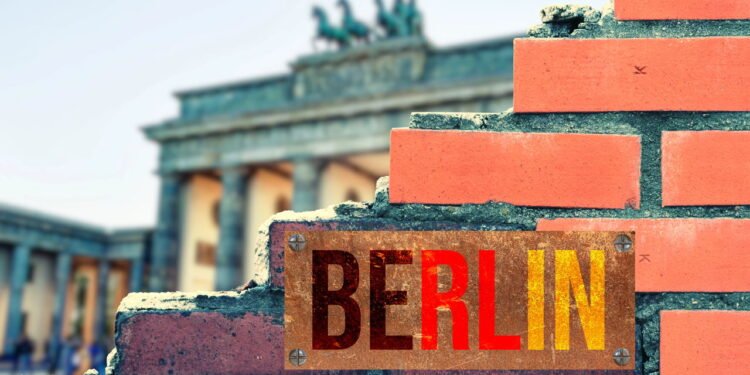 Inscrição "Berlin" em parede de tijolos em ruínas, com o Portão de Brandemburgo desfocado ao fundo - depositphotos.com / stadtratte