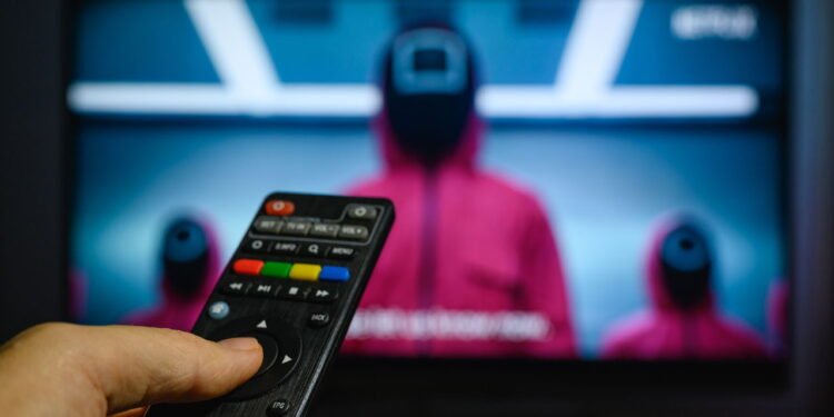 Mão segurando um controle remoto preto em frente a uma TV exibindo uma cena da série coreana Round 6 - depositphotos.com / nardi.bk.ru