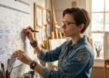 Uma mulher de cabelo curto e óculos, vestindo uma camisa jeans manchada de tinta, escreve com uma caneta preta em um grande calendário de parede branco. O calendário está cheio de notas e prazos manuscritos. Ela está em um estúdio de arte iluminado pela luz natural de uma janela, cercada por cavaletes, telas, pincéis e outros materiais artísticos. Seu olhar está concentrado no calendário.