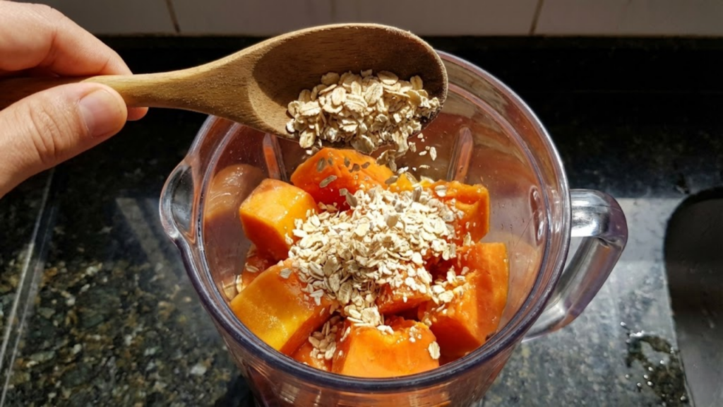 Mão adicionando aveia ao liquidificador com pedaços suculentos de mamão