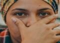 Close-up do rosto de uma pessoa com um lenço estampado na cabeça, cobrindo o nariz e a boca com a mão em um gesto pensativo, focando em seus olhos expressivos.