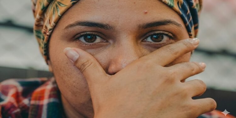 Close-up do rosto de uma pessoa com um lenço estampado na cabeça, cobrindo o nariz e a boca com a mão em um gesto pensativo, focando em seus olhos expressivos.
