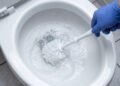 Pessoa usando luva de borracha azul esfrega a parte interna de um vaso sanitário branco com uma escova, enquanto a espuma do produto de limpeza se forma na água