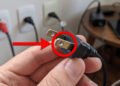 Uma imagem em close-up de uma mão humana segurando um plugue de tomada de estilo americano (tipo A com pinos chatos), com um círculo vermelho e uma seta vermelha apontando para os orifícios nos pinos de metal. No fundo, fora de foco, há tomadas elétricas de estilo brasileiro (tipo N) com plugues inseridos na parede e eletrônicos sobre uma mesa.