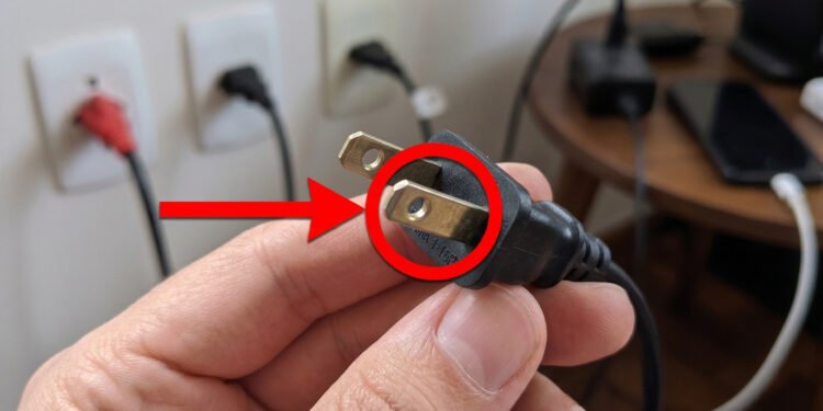 Uma imagem em close-up de uma mão humana segurando um plugue de tomada de estilo americano (tipo A com pinos chatos), com um círculo vermelho e uma seta vermelha apontando para os orifícios nos pinos de metal. No fundo, fora de foco, há tomadas elétricas de estilo brasileiro (tipo N) com plugues inseridos na parede e eletrônicos sobre uma mesa.