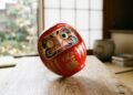 Boneco Daruma vermelho japonês inclinado voltando à posição vertical sobre mesa de madeira com luz suave