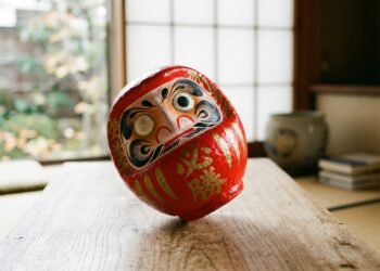 Boneco Daruma vermelho japonês inclinado voltando à posição vertical sobre mesa de madeira com luz suave