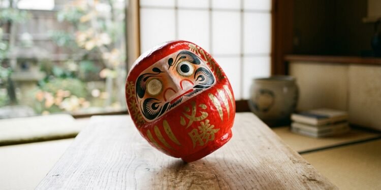 Boneco Daruma vermelho japonês inclinado voltando à posição vertical sobre mesa de madeira com luz suave