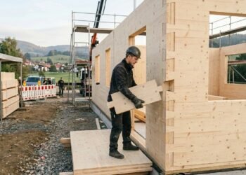 Paredes de casa em construção com blocos de madeira maciça encaixados sem cimento, com trabalhador montando bloco com as mãos