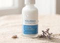 Frasco de produto pós-praia multifuncional sobre areia clara, com gotas de água, ao lado de uma pequena pedra e flores secas, destacando a hidratação da pele.