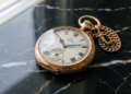 Relógio de bolso antigo da marca Omega em ouro, com corrente, posicionado sobre uma superfície de mármore preto com veios brancos, destacando a elegância clássica.