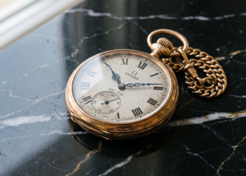 Relógio de bolso antigo da marca Omega em ouro, com corrente, posicionado sobre uma superfície de mármore preto com veios brancos, destacando a elegância clássica.