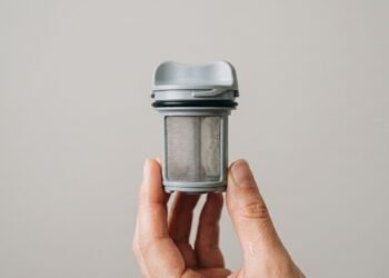 Mão segurando um filtro de máquina de lavar cinza, composto por uma estrutura de plástico e uma malha fina de metal, isolado em fundo neutro.