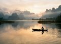 Lago tranquilo ao amanhecer com barco de pesca e montanhas enevoadas em paisagem chinesa