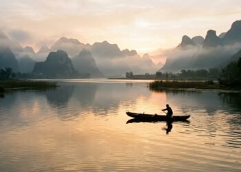 Lago tranquilo ao amanhecer com barco de pesca e montanhas enevoadas em paisagem chinesa