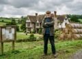 Homem de braços cruzados diante de mansão estilo castelo Tudor parcialmente revelada por trás de fardos de feno, no campo inglês.