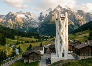 Torre branca impressa em 3D de 30 metros erguendo-se acima dos telhados de pequena vila alpina na Suíça com montanhas ao fundo