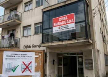 Sacada de apartamento fechada com vidro exibindo adesivo de "Obra Embargada" e aviso de proibição de alteração de fachada em primeiro plano.