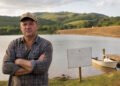 Homem à beira de um grande lago represado em propriedade rural arborizada, com barragem de terra ao fundo.
