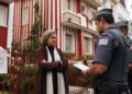 Mulher idosa de braços cruzados diante de sobrado pintado com listras vermelhas e brancas, em rua de casas claras em Londres.