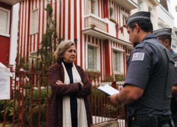 Mulher idosa de braços cruzados diante de sobrado pintado com listras vermelhas e brancas, em rua de casas claras em Londres.