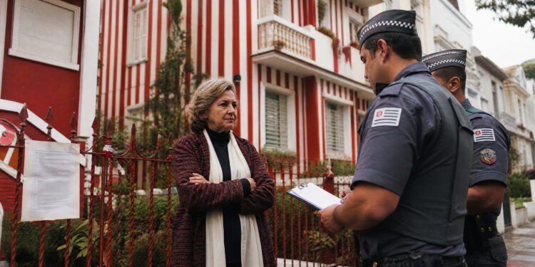 Mulher idosa de braços cruzados diante de sobrado pintado com listras vermelhas e brancas, em rua de casas claras em Londres.