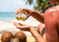 Mão derramando óleo de coco na praia com ombro queimado de sol ao fundo, mostrando o risco de usar o óleo como bronzeador