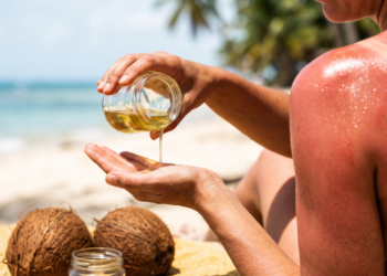 Mão derramando óleo de coco na praia com ombro queimado de sol ao fundo, mostrando o risco de usar o óleo como bronzeador