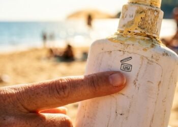 Mão segurando frasco de protetor solar vencido com creme amarelado e textura separada, com ombro queimado de sol ao fundo na praia