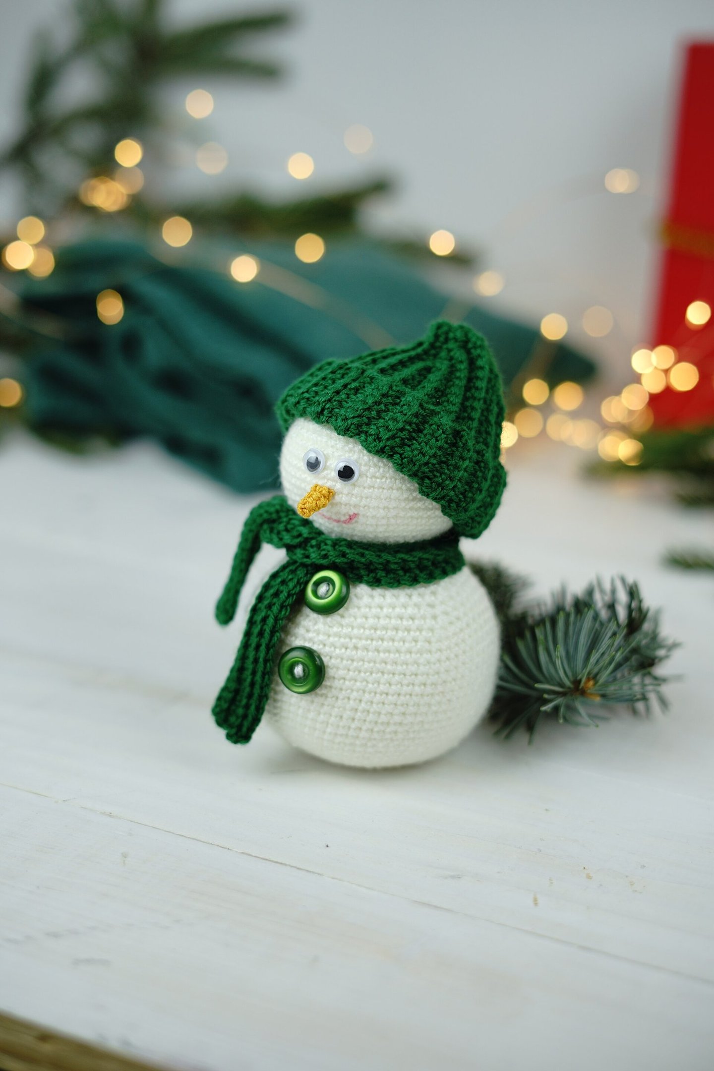 Boneco de neve de crochê branco com gorro e cachecol verdes, botões verdes e nariz laranja, sobre uma mesa de madeira branca.